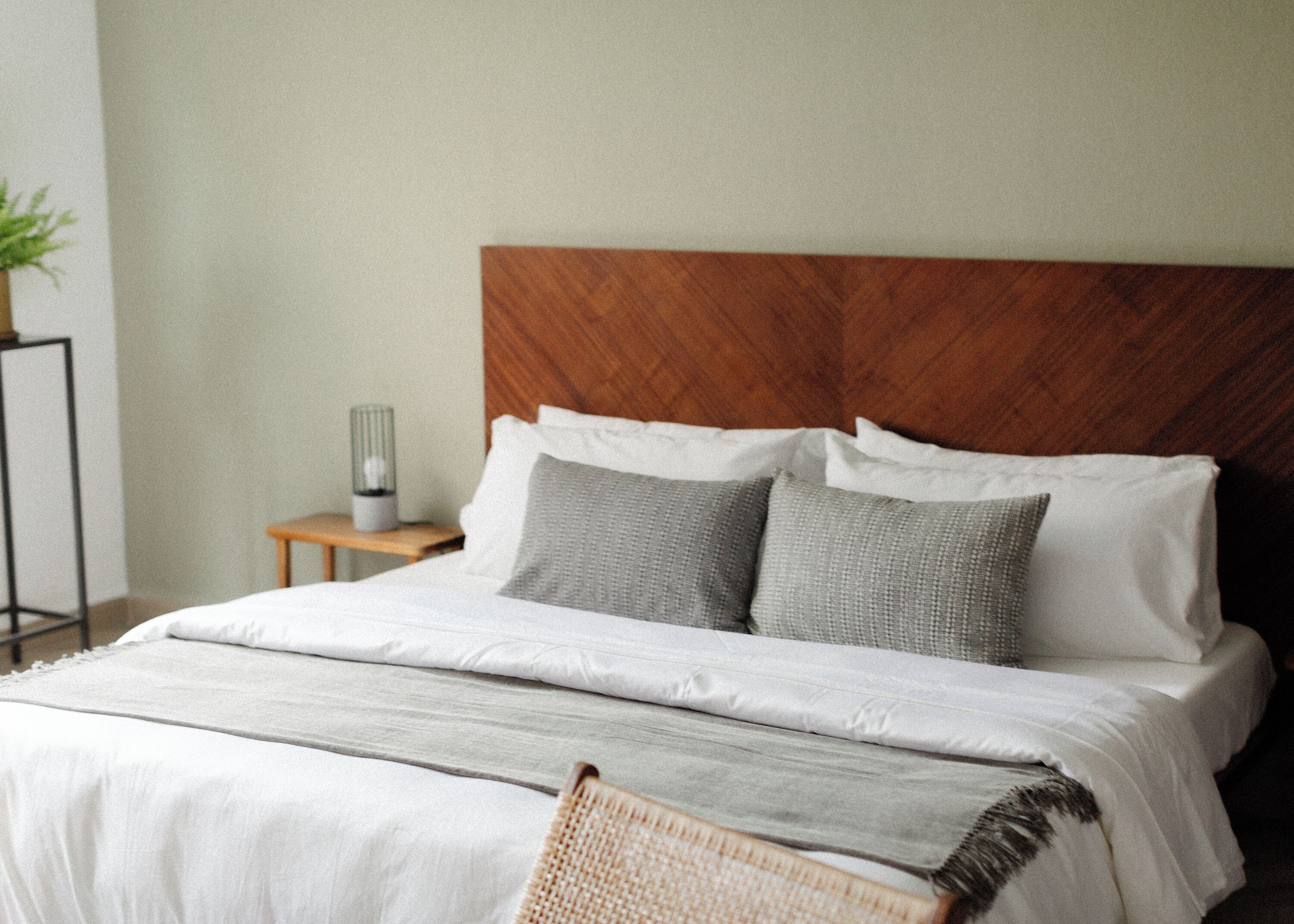 Una cama bien hecha con cabecera de madera, una mesita de noche con lámpara y una silla de mimbre.
