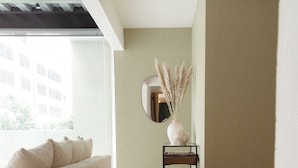 Ein modernes Wohnzimmer mit einer Couch, einem runden gewebten Hocker, einem kleinen Beistelltisch mit einer Vase mit Pampasgras und einem großen Spiegel an der Wand.