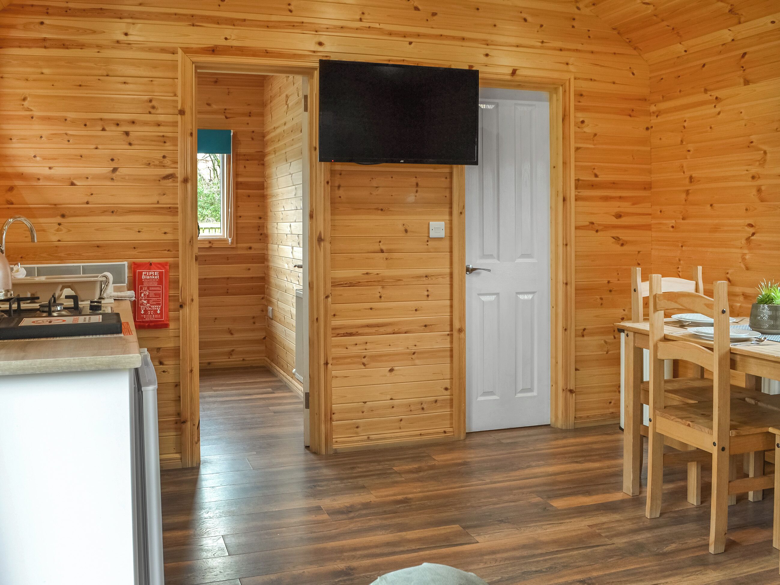 A wooden cabin interior with a kitchen area, a dining table with chairs, and a television mounted on the wall.