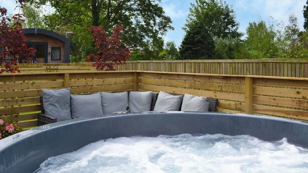 Un bain à remous doté d’une terrasse en bois et de coussins, entouré d’une clôture en bois et d’arbres.