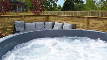 A hot tub with a wooden deck and cushions, surrounded by a wooden fence and trees.