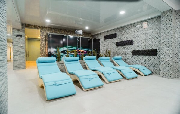 A spa room with multiple sun loungers, a large window, and a wall-mounted screen displaying a relaxing scene.