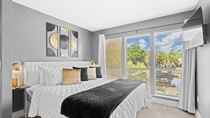 Une chambre moderne avec un grand lit, une table de chevet, une lampe et une vue sur la verdure par la fenêtre.