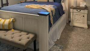 Ein Schlafzimmer mit Bett, Nachttischen, einer Bank, einem Fenster mit Vorhängen und einer Deckenleuchte.