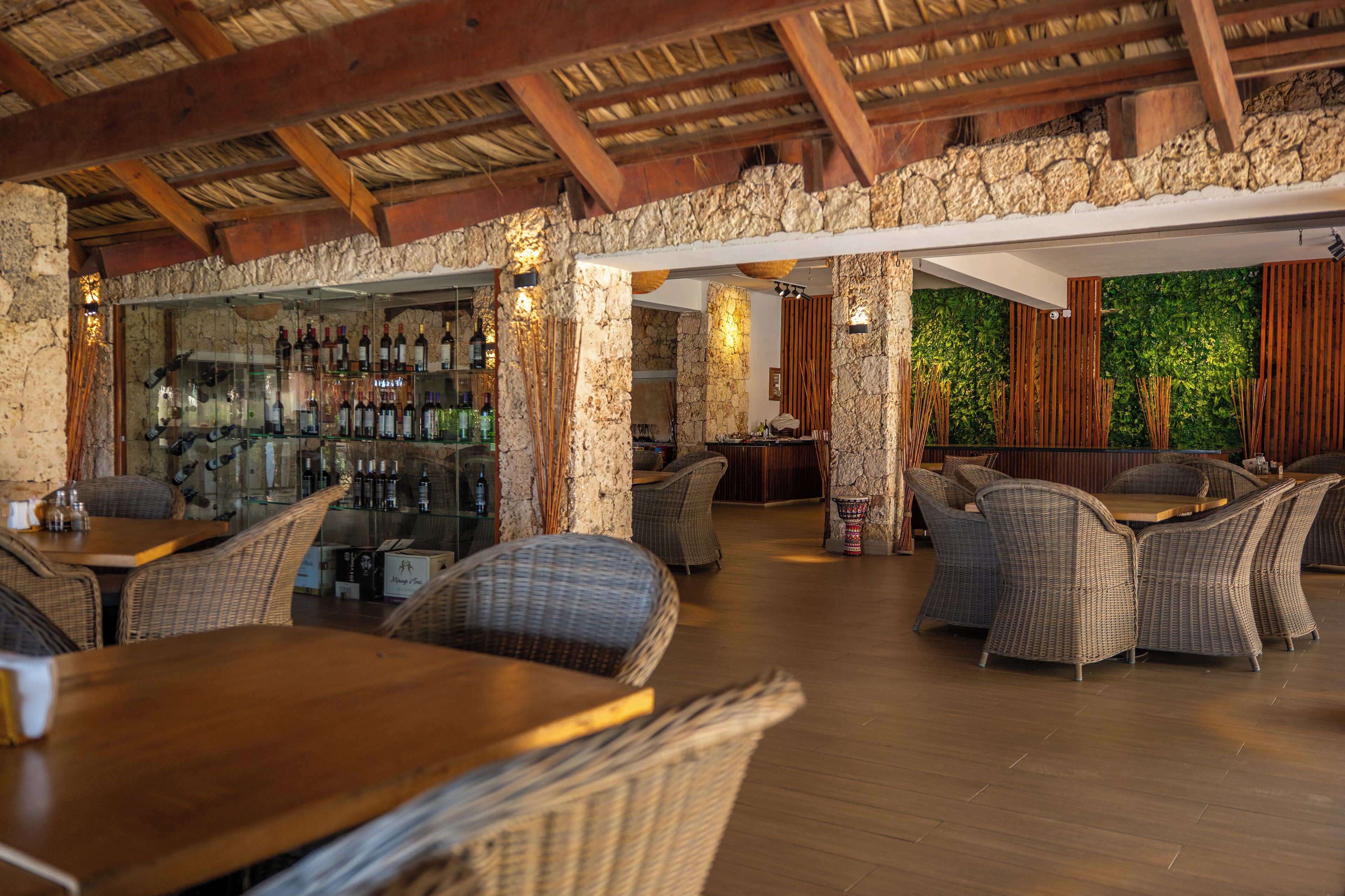 A restaurant with wicker furniture, a bar area with bottles, and a stone wall backdrop.