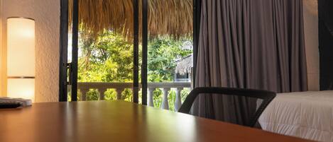 Habitación de hotel con una mesa de madera, una silla, una cama con ropa de cama blanca, un ventanal con cortinas y vistas al exterior con vegetación.