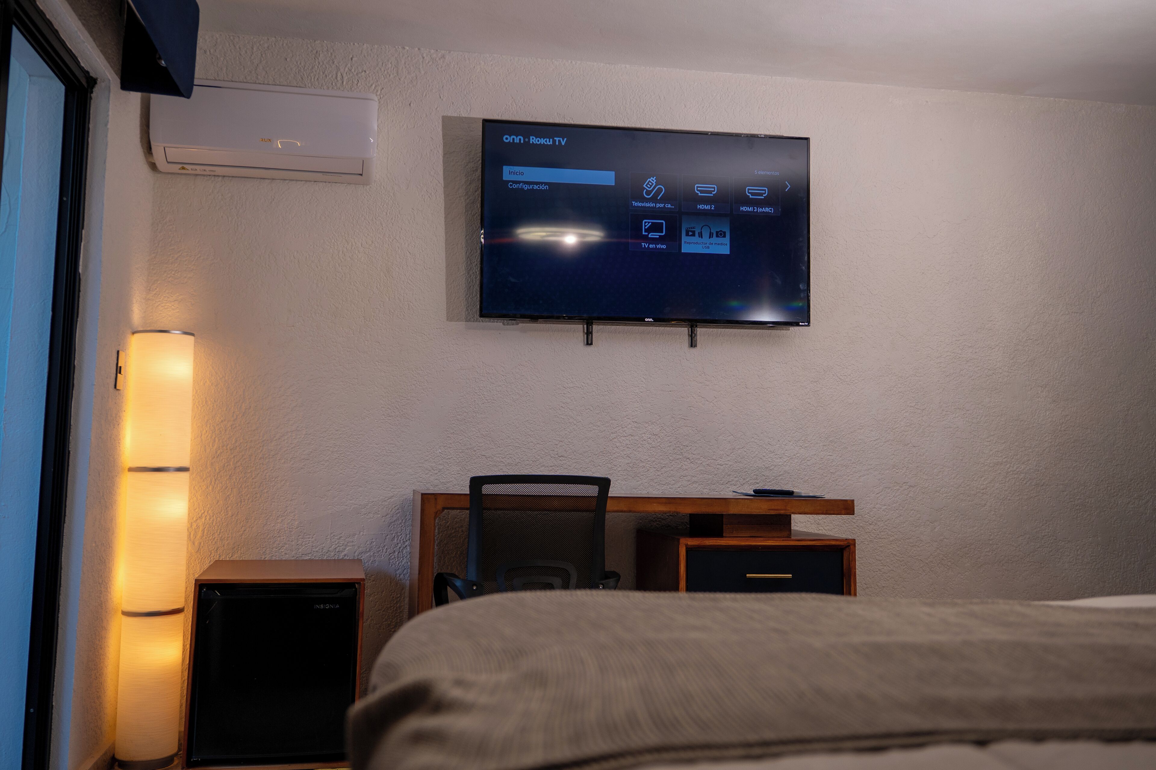 A hotel room with a flat-screen TV mounted on the wall, a desk with a chair, a bed, and a standing speaker.