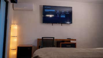 A hotel room with a flat-screen TV mounted on the wall, a desk with a chair, a bed, and a standing speaker.