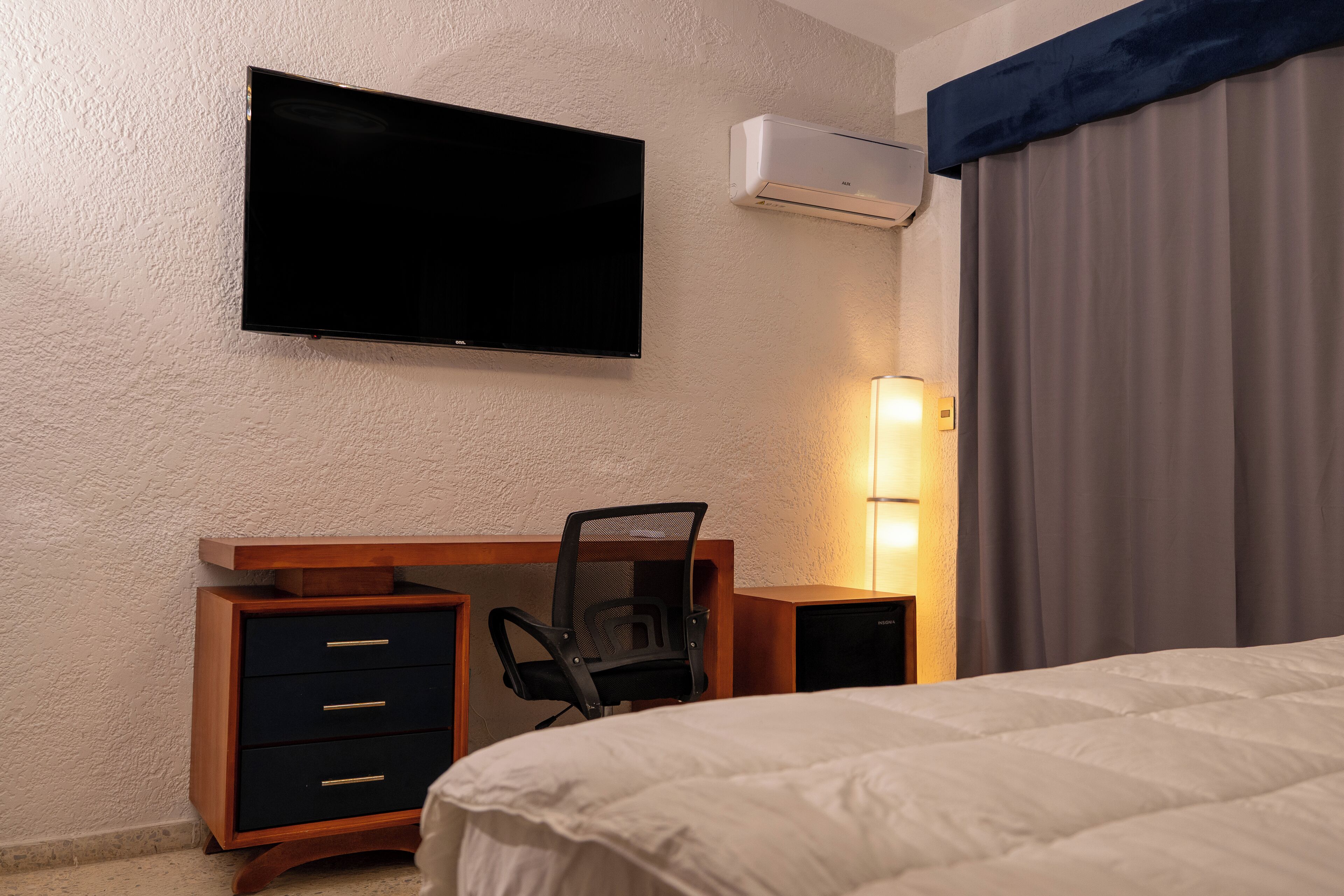 A hotel room with a bed, a desk with a chair, a television, an air conditioning unit, and a lamp.