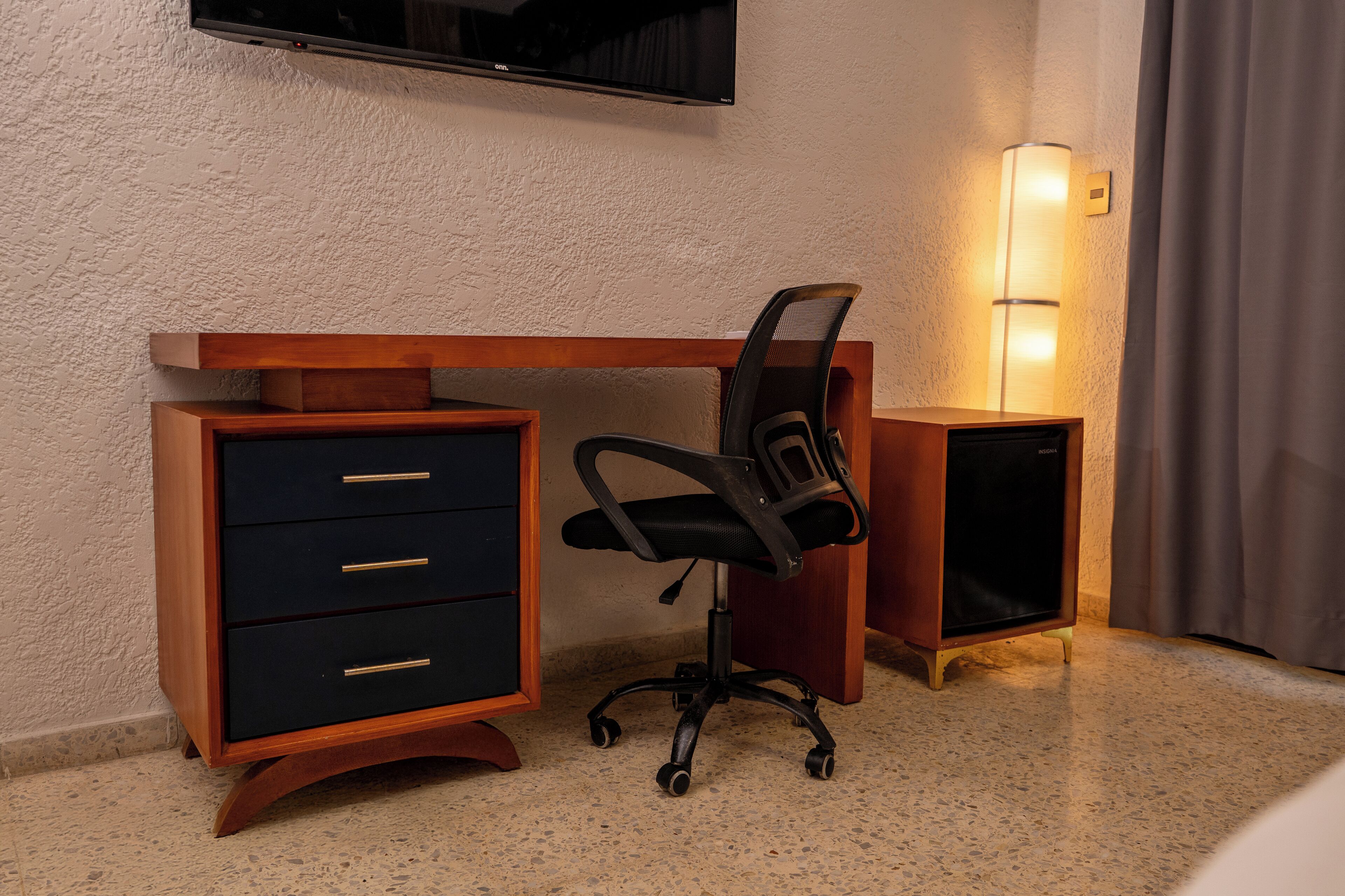 A hotel room with a wooden desk, a black office chair, a TV mounted on the wall, and a bedside lamp.