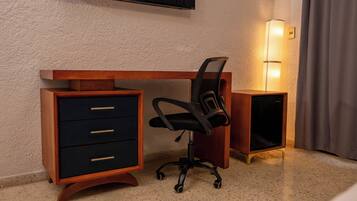 A hotel room with a wooden desk, a black office chair, a TV mounted on the wall, and a bedside lamp.