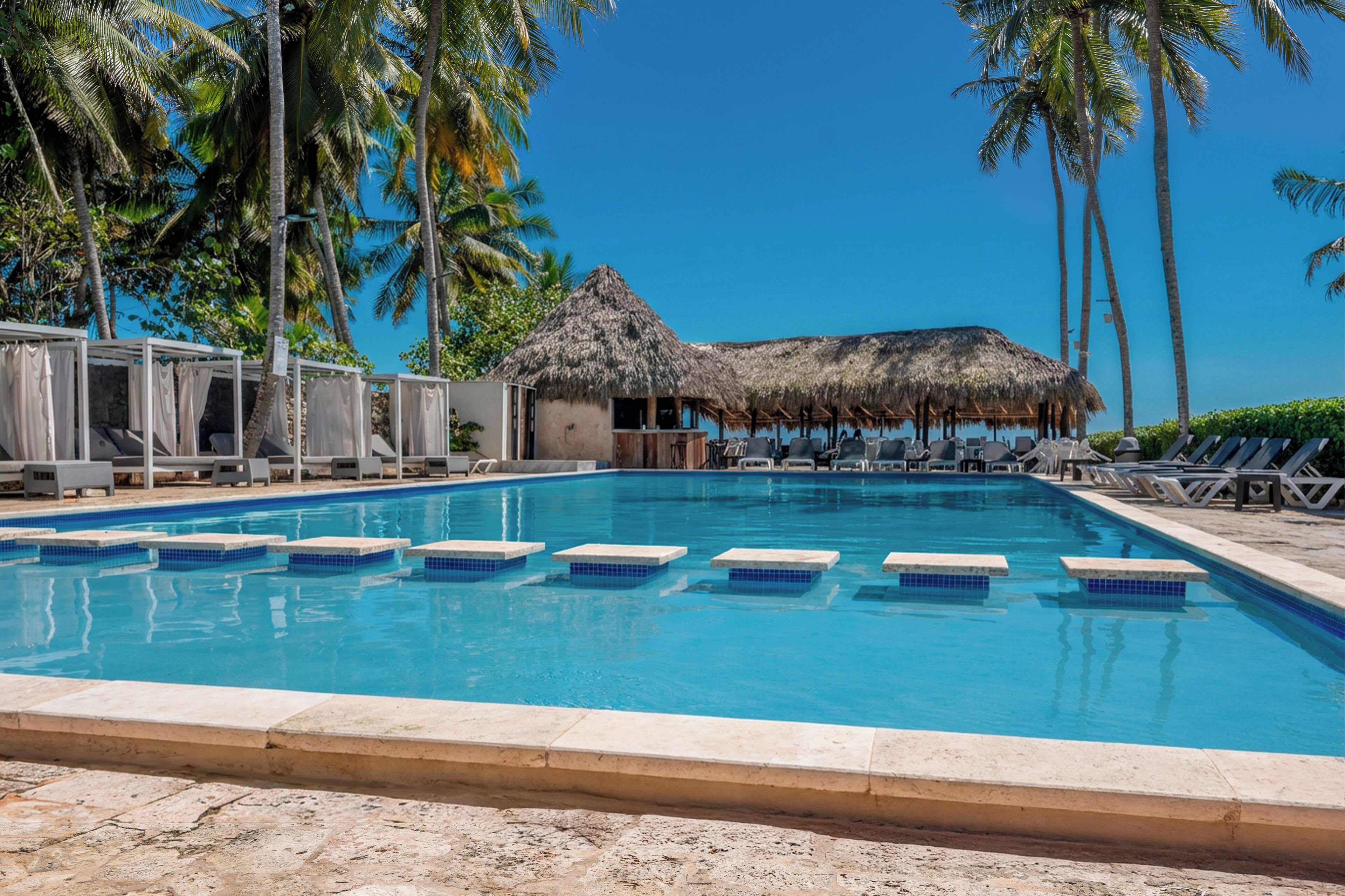 A swimming pool with lounge chairs and a thatched-roof structure, surrounded by palm trees.