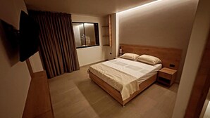 A modern bedroom with a wooden bed, a bedside table, a TV mounted on the wall, and a window with curtains. - Hotel La Casona San Sebastian  (Puerto Triunfo)