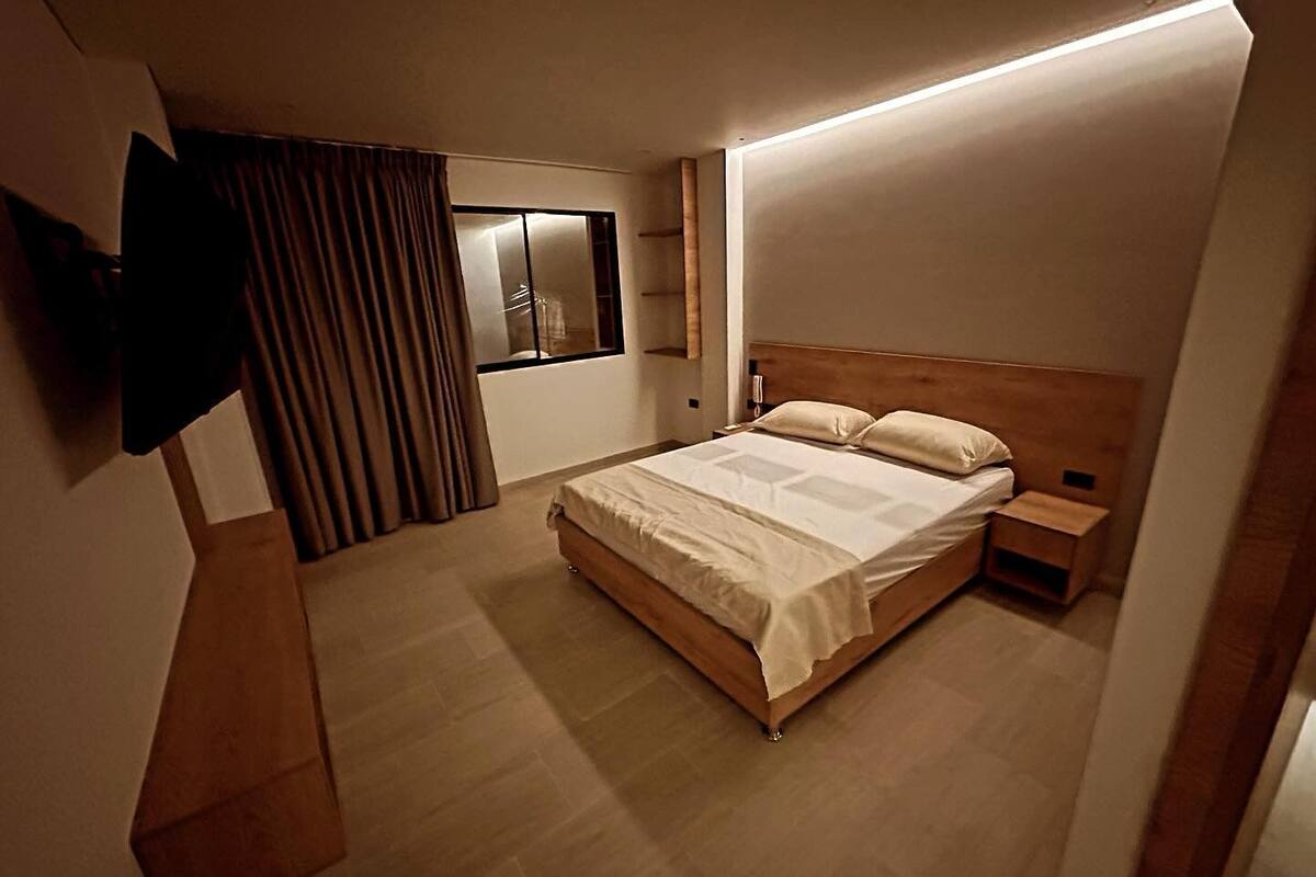 Une chambre moderne avec un lit en bois, une table de chevet, un téléviseur fixé au mur et une fenêtre avec des rideaux.
