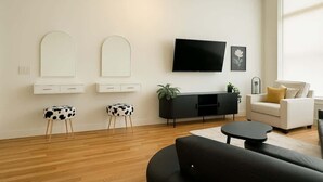 Un salon moderne avec un canapé noir, un fauteuil blanc, une table basse noire et un téléviseur à écran plat fixé au mur. On y trouve deux meubles blancs avec des miroirs et un meuble noir pour équipements audiovisuels agrémenté d’une plante.