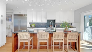 A modern kitchen with white cabinets, a stainless steel refrigerator, a central island with bar stools, and a large window providing natural light. - Scenic 4BR/3BA Waterfront Getaway with private dock, kayaks & Stunning Views (Dartmouth)