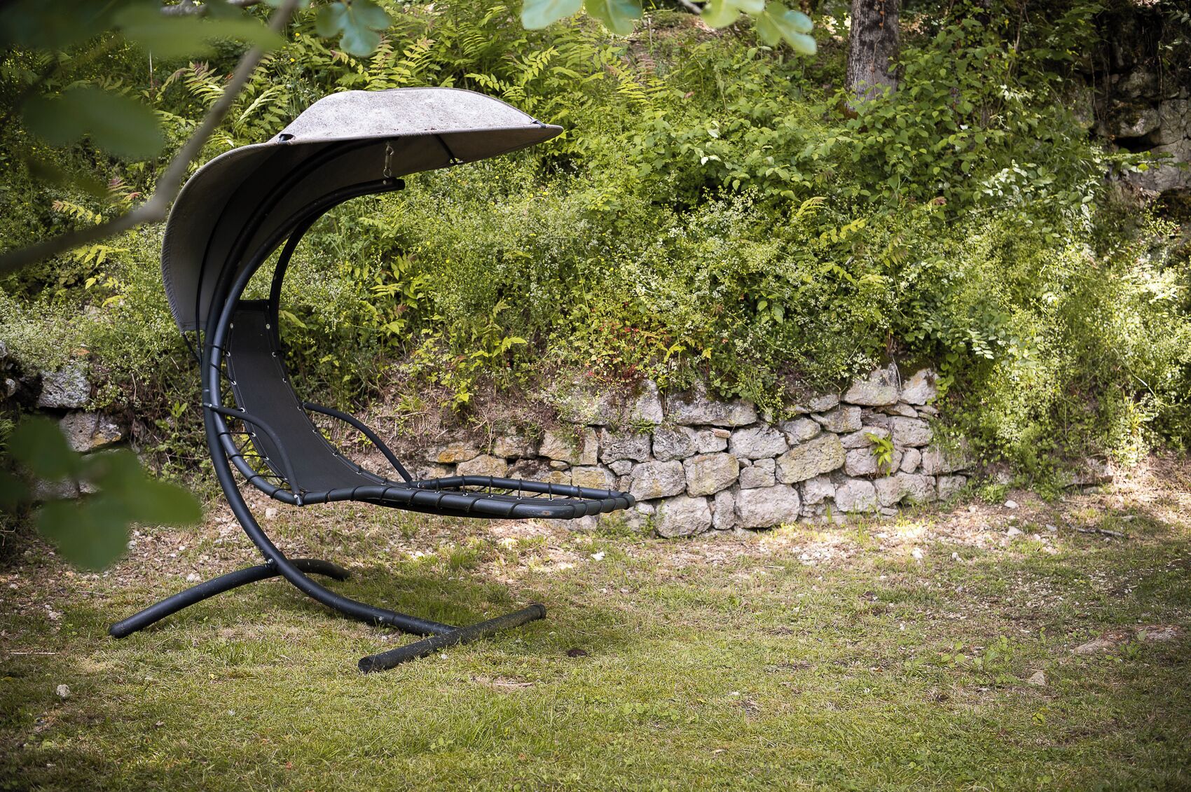 Una silla de jardín con un diseño original, ubicada sobre césped, con un muro de piedra y abundante follaje al fondo.