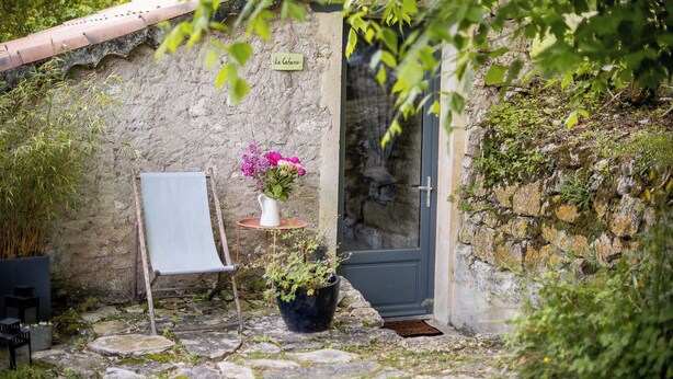 A rustic stone wall with a door, a small table with a vase of flowers, a chair, and a potted plant.