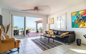 Un salon moderne avec un canapé, une table basse et des fauteuils, une grande fenêtre donnant sur la mer et un ventilateur de plafond.