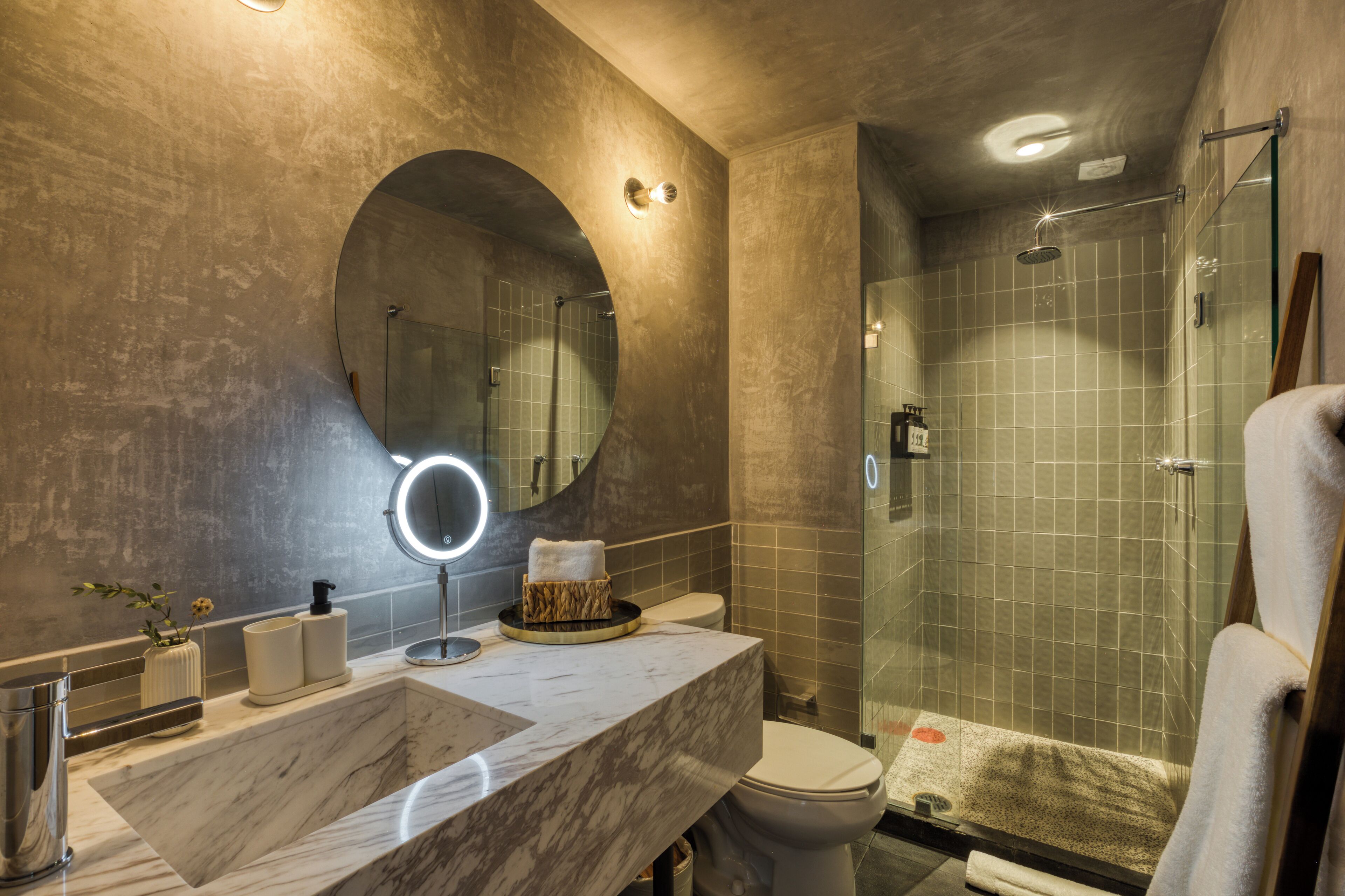 Een badkamer met een marmeren wastafel, een ronde spiegel met verlichting, een toilet en een douchecabine met glazen wanden.