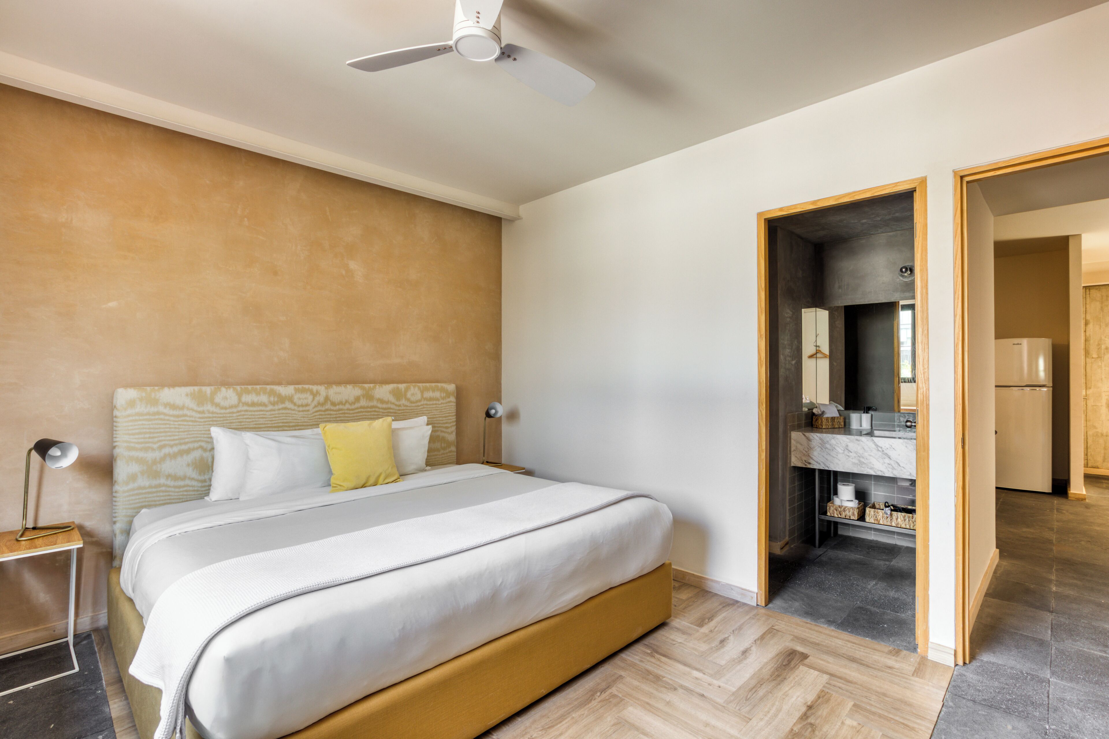 A modern hotel room with a large bed, a ceiling fan, a wooden headboard, and a view into the bathroom.