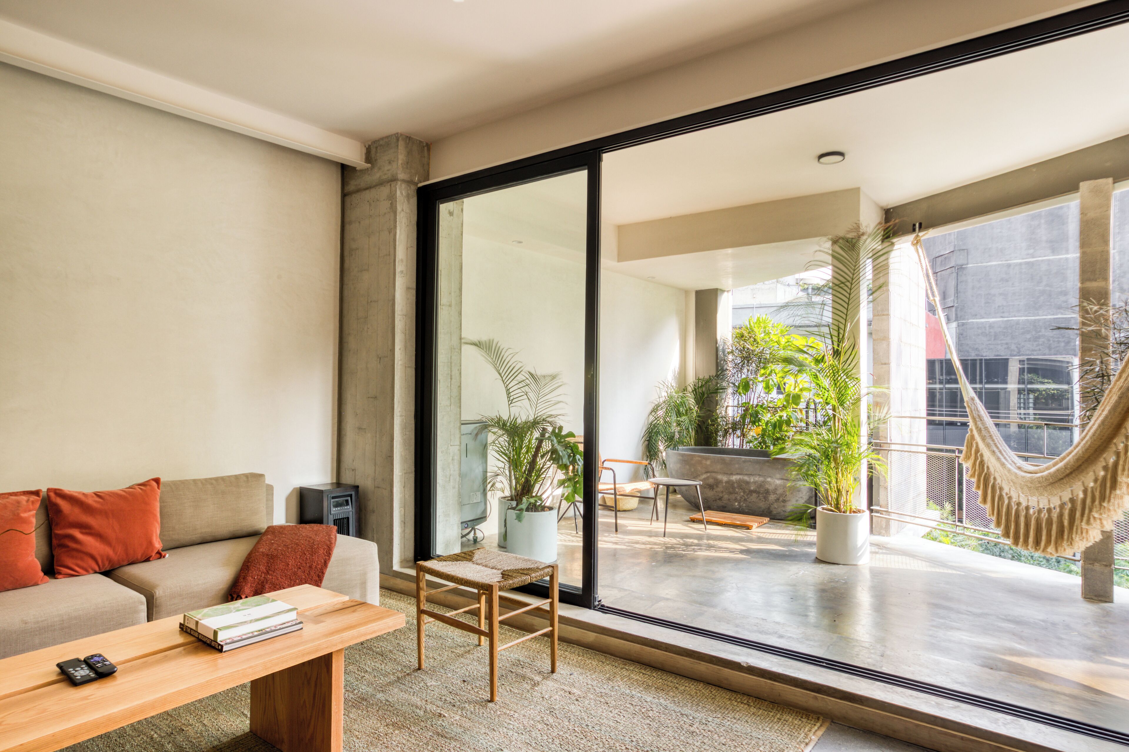 A modern living room with a sofa, coffee table, and a large sliding glass door leading to an outdoor patio area with plants and a hammock.