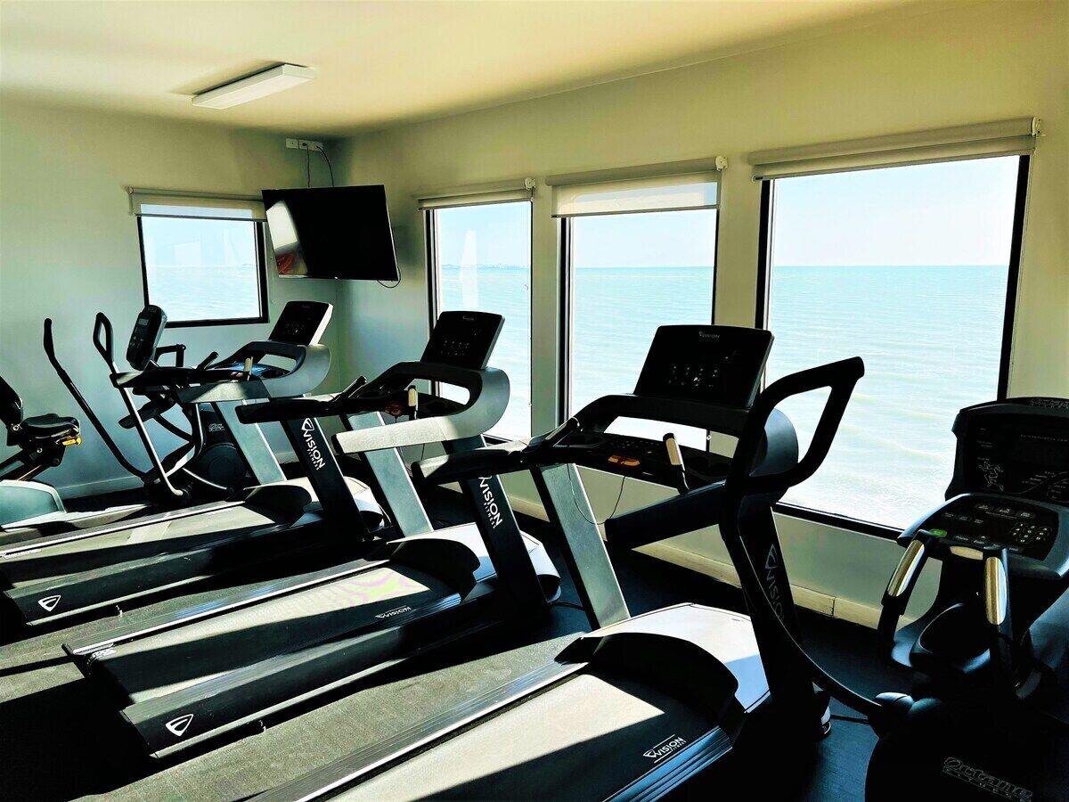 A gym with treadmills and exercise bikes, large windows with a view of the sea, and a flat-screen TV mounted on the wall.