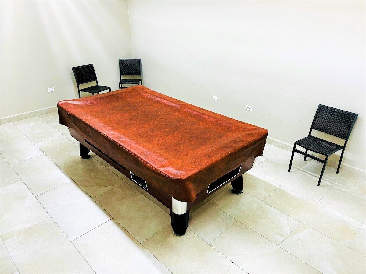 Una mesa de billar rectangular con superficie de fieltro rojo, rodeada por dos sillas negras de respaldo trenzado.