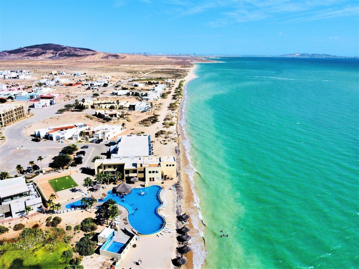 An aerial view of a coastal resort with a large swimming pool, beach, and several buildings.