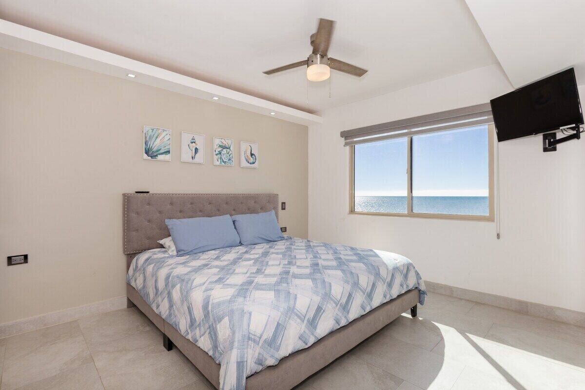 A bedroom with a bed, ceiling fan, artwork, window, and TV.