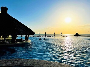 A serene pool area with a thatched-roof structure, calm water, and a sunset in the background.