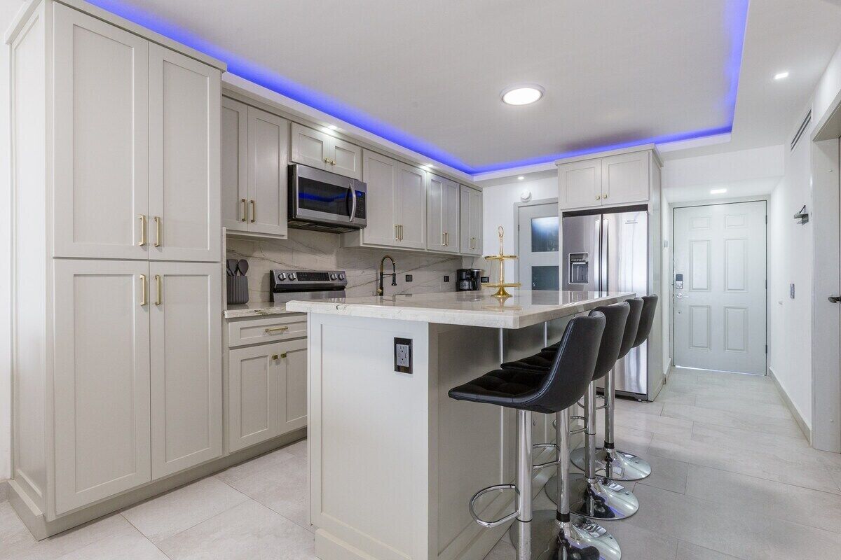 A modern kitchen with white cabinets, a central island with bar stools, stainless steel appliances, and blue LED lighting.
