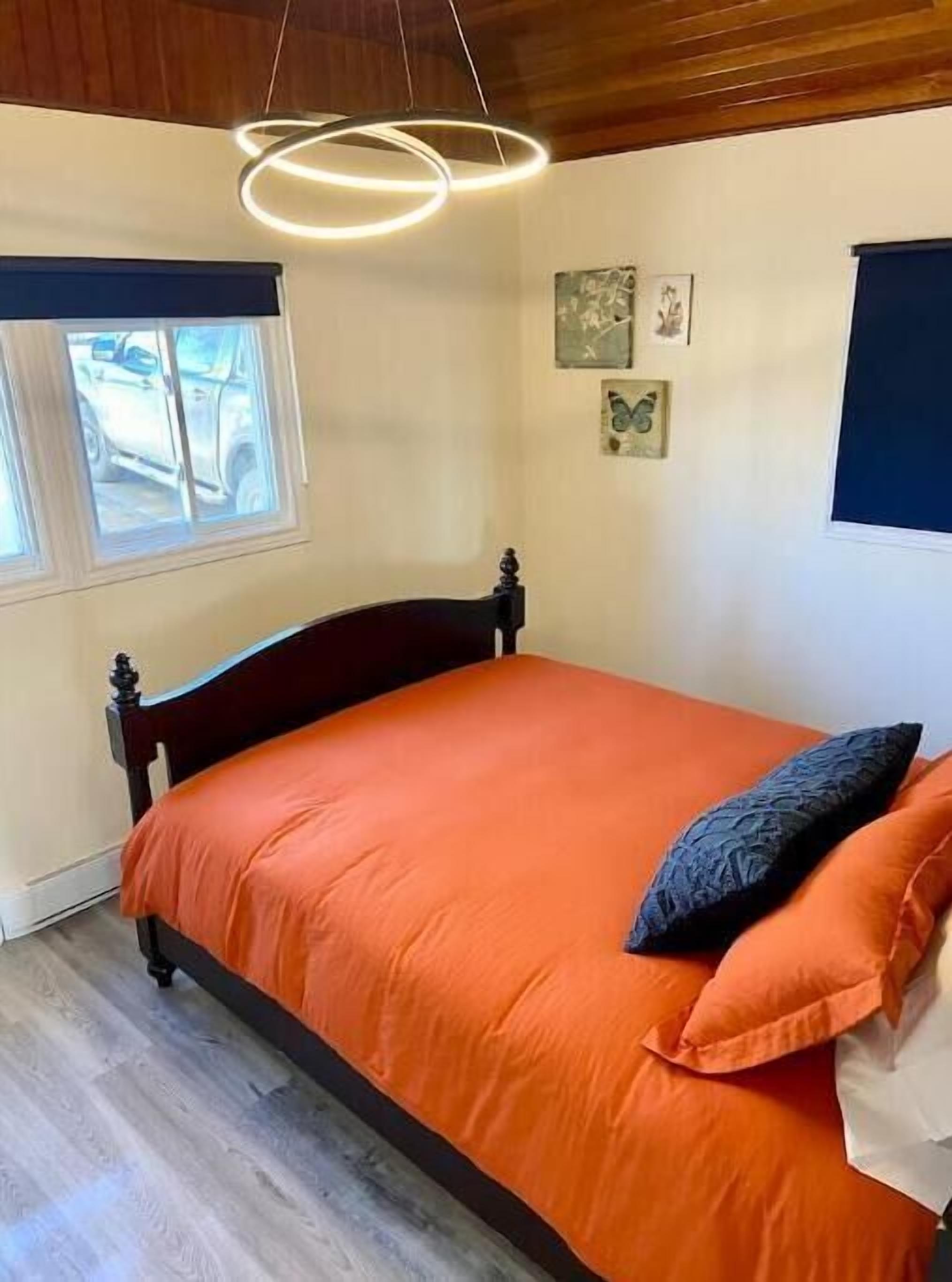 Ein Schlafzimmer mit einem Holz-Kopfteil, einer orangefarbenen Bettdecke, einem dunklen Kissen und einem Fenster mit Blick auf ein Auto.