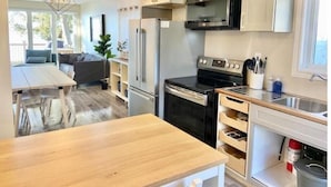 A compact kitchen with white cabinets, a stainless steel refrigerator, an oven, and a microwave. There is a wooden table and a built-in sink with a window above it. - Lakeside Cottage Getaway South Shore Lake Scugog

 (Caesarea)