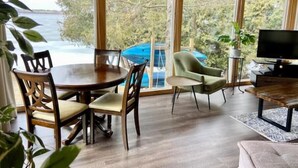 A room with a wooden dining table and chairs, a television, and a view of the sea through large windows. - Cozy Lakeside Cottage on Lake Scugog

 (Little Britain)