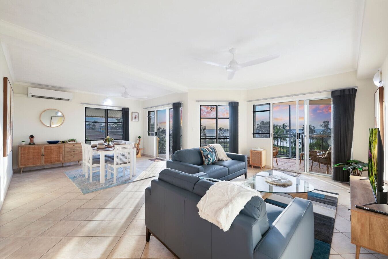 A spacious living room with a grey sofa, a coffee table, and a dining area with a table and chairs. There is a balcony with a view of the ocean.