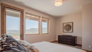 A bedroom with a large bed, a dresser, and a view of mountains through the windows.