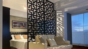 A modern bedroom with a bed, a sofa, a decorative black and white wall panel, and a large window with a view.