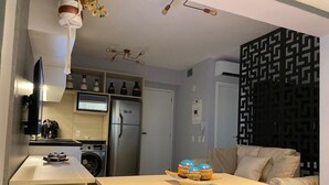 A modern kitchen with a built-in oven, a coffee machine, and a washing machine. There is a dining table with a decorative bowl and a sofa in the living area.