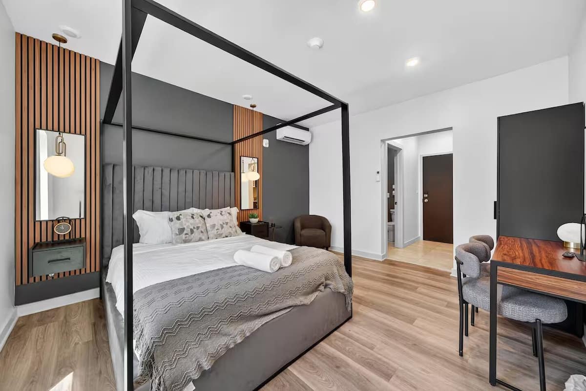Quarto moderno com cama de dossel, escrivaninha com cadeira e banheiro visível por uma porta aberta.