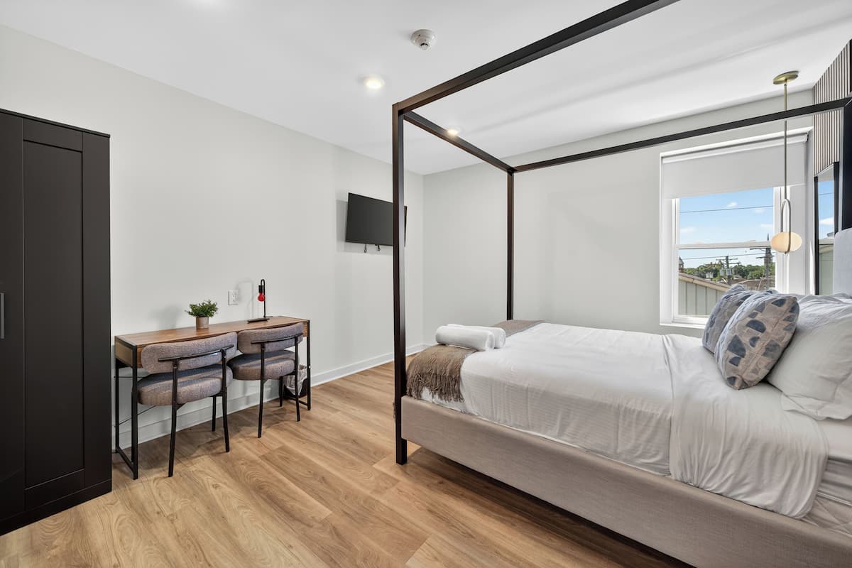 Quarto com cama de dossel, uma escrivaninha com cadeiras, TV fixada na parede e janela com vista.