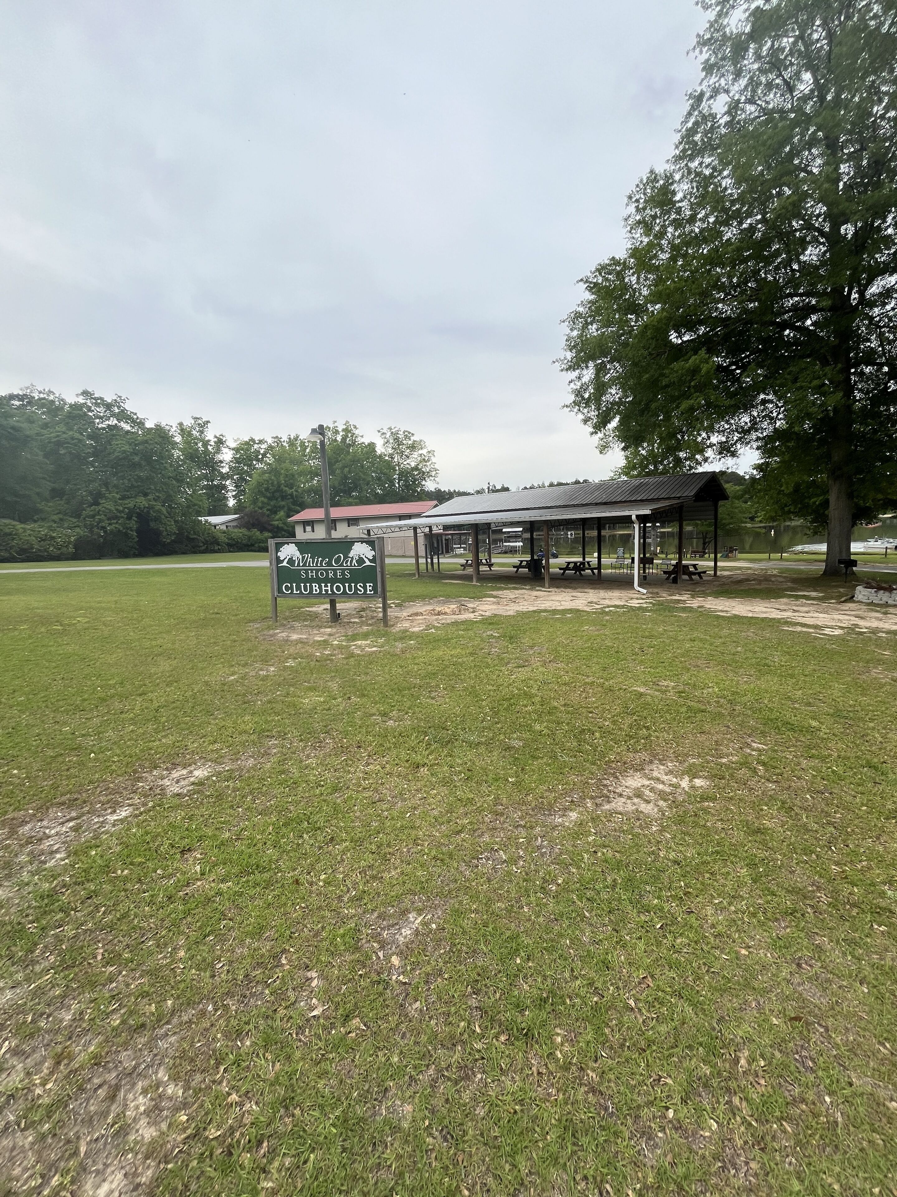 Un espacio verde con un letrero que indica Club House de White Oak Shores, un pabellón cubierto con mesas de picnic y un edificio de techo rojo al fondo.