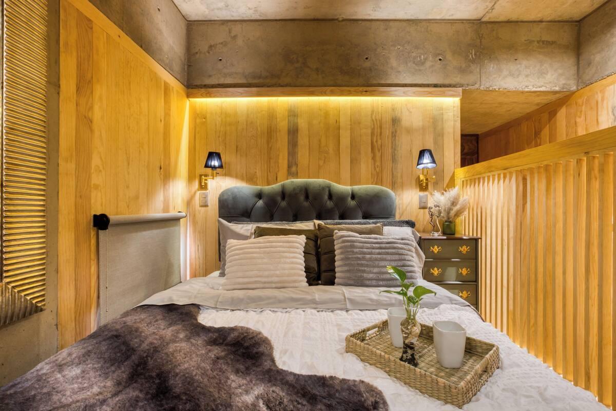 Un dormitorio con cabecera capitonada, paneles de madera, mesita de noche con lámparas, cómoda y planta en la mesita.