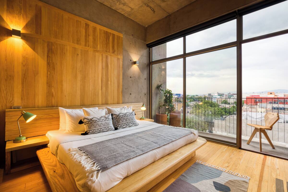 Un dormitorio moderno con una cama grande, cabecera de madera y vistas a la ciudad a través de amplios ventanales.
