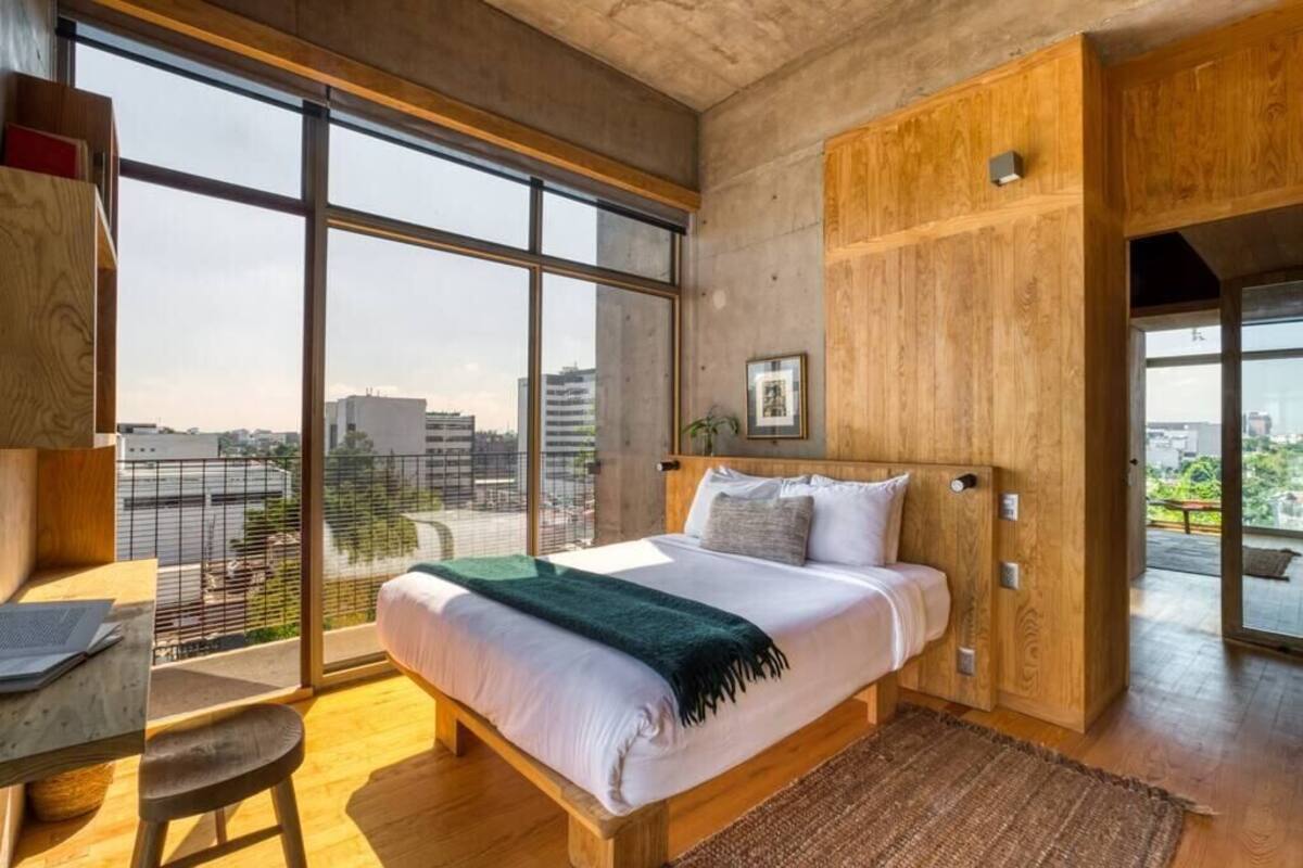 Un dormitorio moderno con una cama grande, cabecero de madera y vistas a la ciudad a través de amplias ventanas.