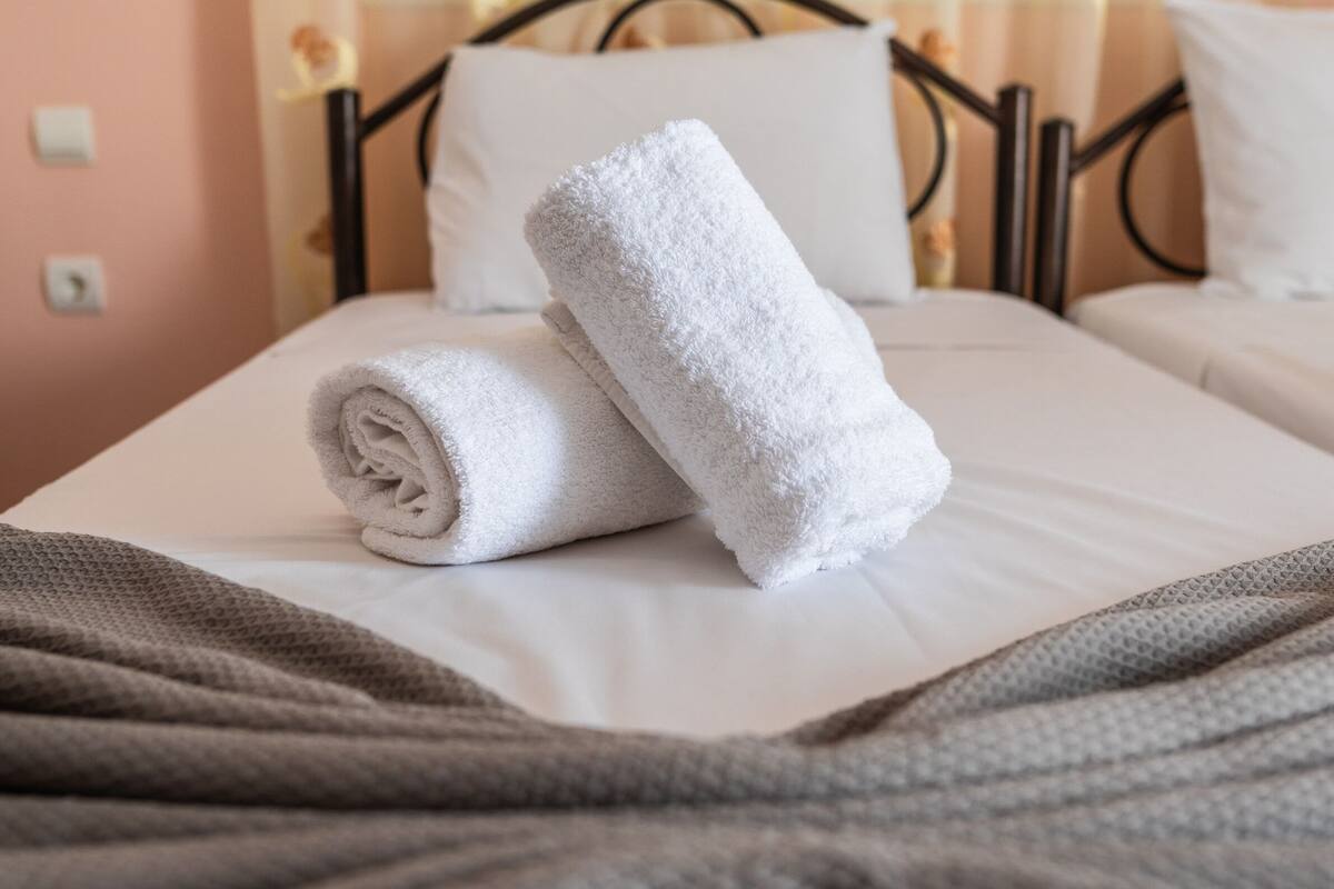 A neatly made bed with white linens and rolled towels, a brown blanket, and a window with floral curtains.
