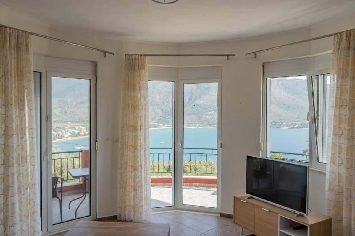 A room with a TV, a balcony with a table and chairs, and large windows offering a view of a lake and mountains.