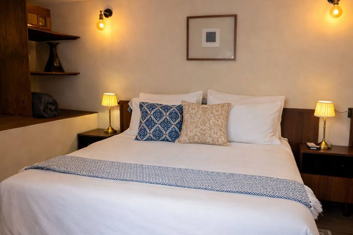 Una cama bien hecha con sábanas blancas y un edredón estampado, dos lámparas de noche, un cuadro en la pared y una mesita de noche de madera con una lámpara.
