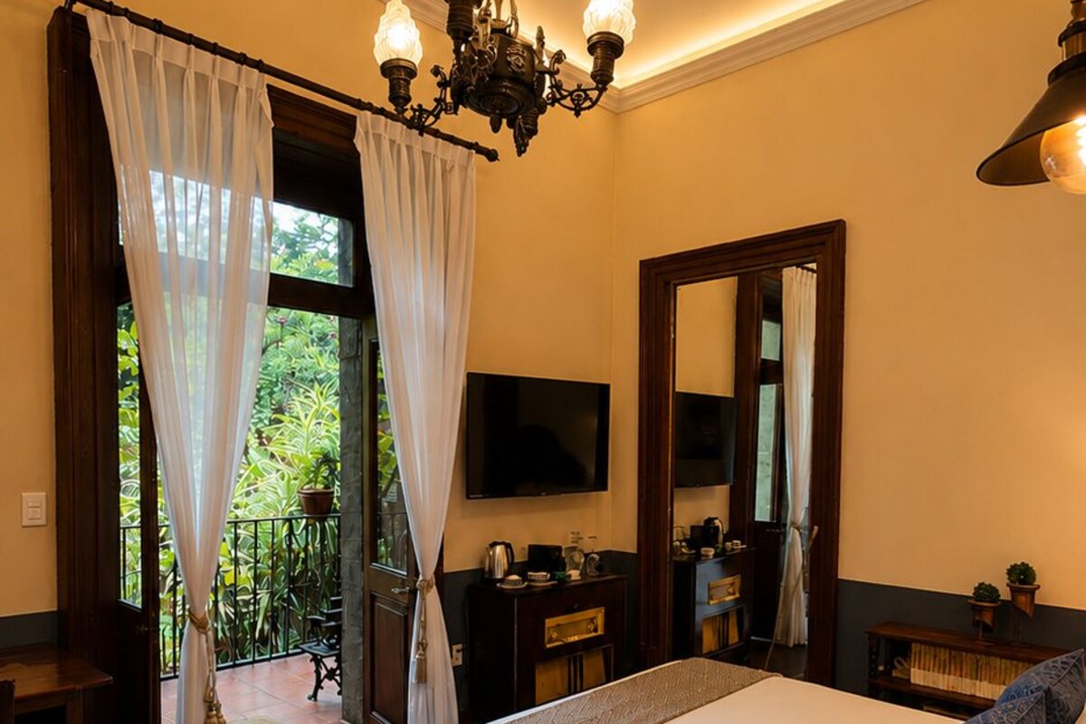Une chambre d’hôtel avec un grand lit, une télévision, un lustre, un miroir et une vue sur la verdure environnante.