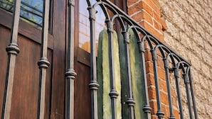 Un balcón con barandilla de hierro forjado y una puerta de madera, frente a un muro de ladrillo.